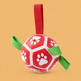 Red and white paw-print rope ball dog toy with red strap and green loops on a warm peach background